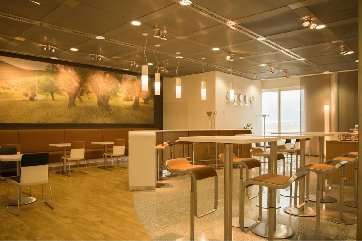 Relaxed and airy seating area at the Lufthansa Business Lounge at Athens Airport, featuring a scenic mural of an olive grove on the back wall. The dining section includes modern furniture with high stools and bar chairs.
