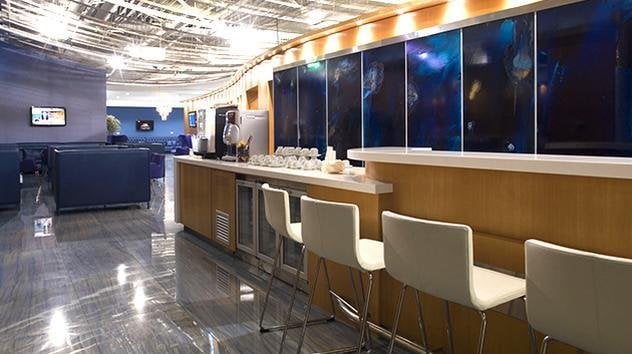 Interior of the Melina Merkouri Lounge at Athens Airport, featuring a contemporary bar setup with bar stools along a polished wooden counter. Behind the counter a series of blue panels enhance the modern ambience. The lounge has plush blue seats.