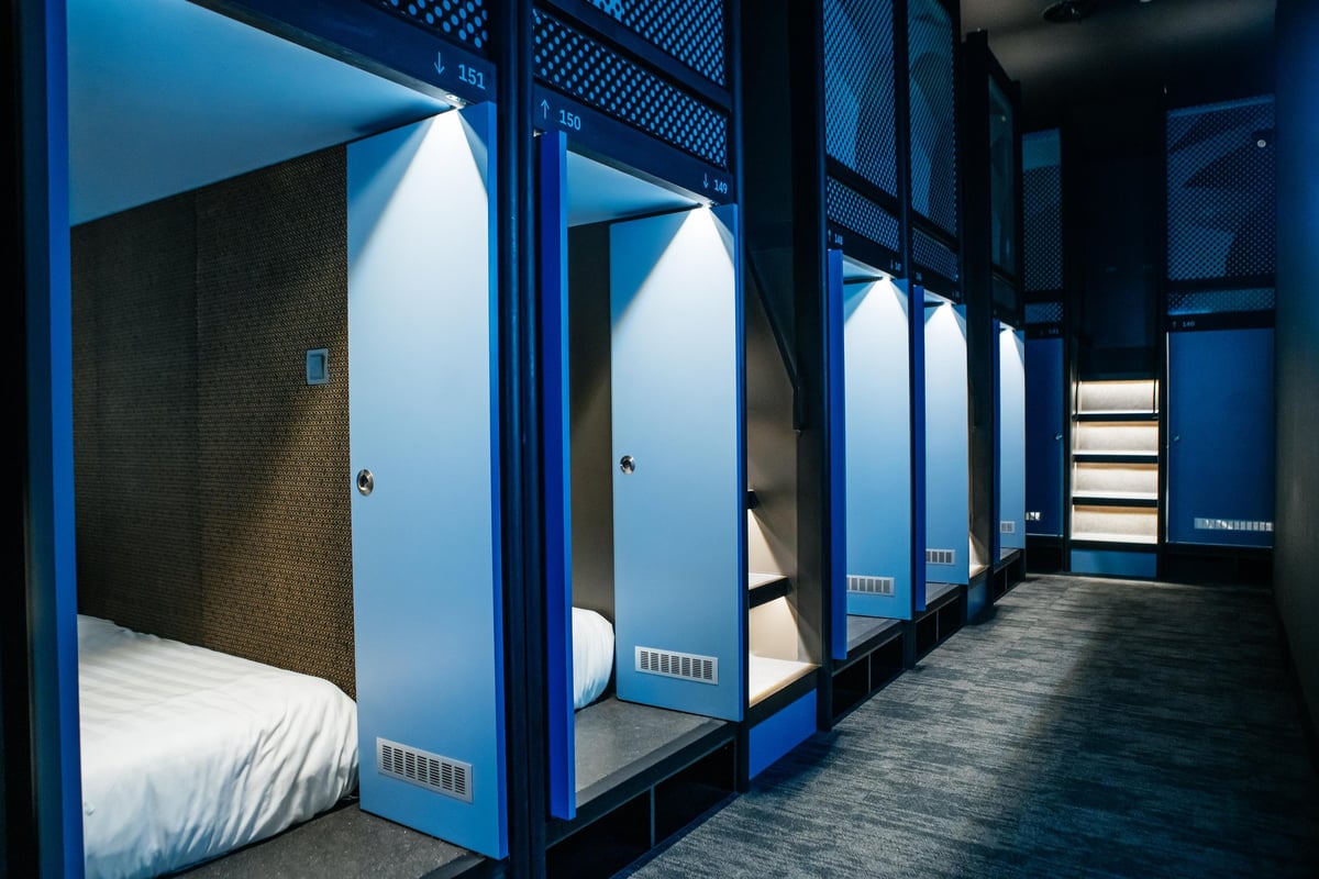 Row of sleek blue sleeping pods at CapsuleTransit KLIA Terminal 2 (Airside), each with a number label and private sliding door, set in a modern, dimly lit corridor with carpeted flooring and a staircase leading to an upper pod level.