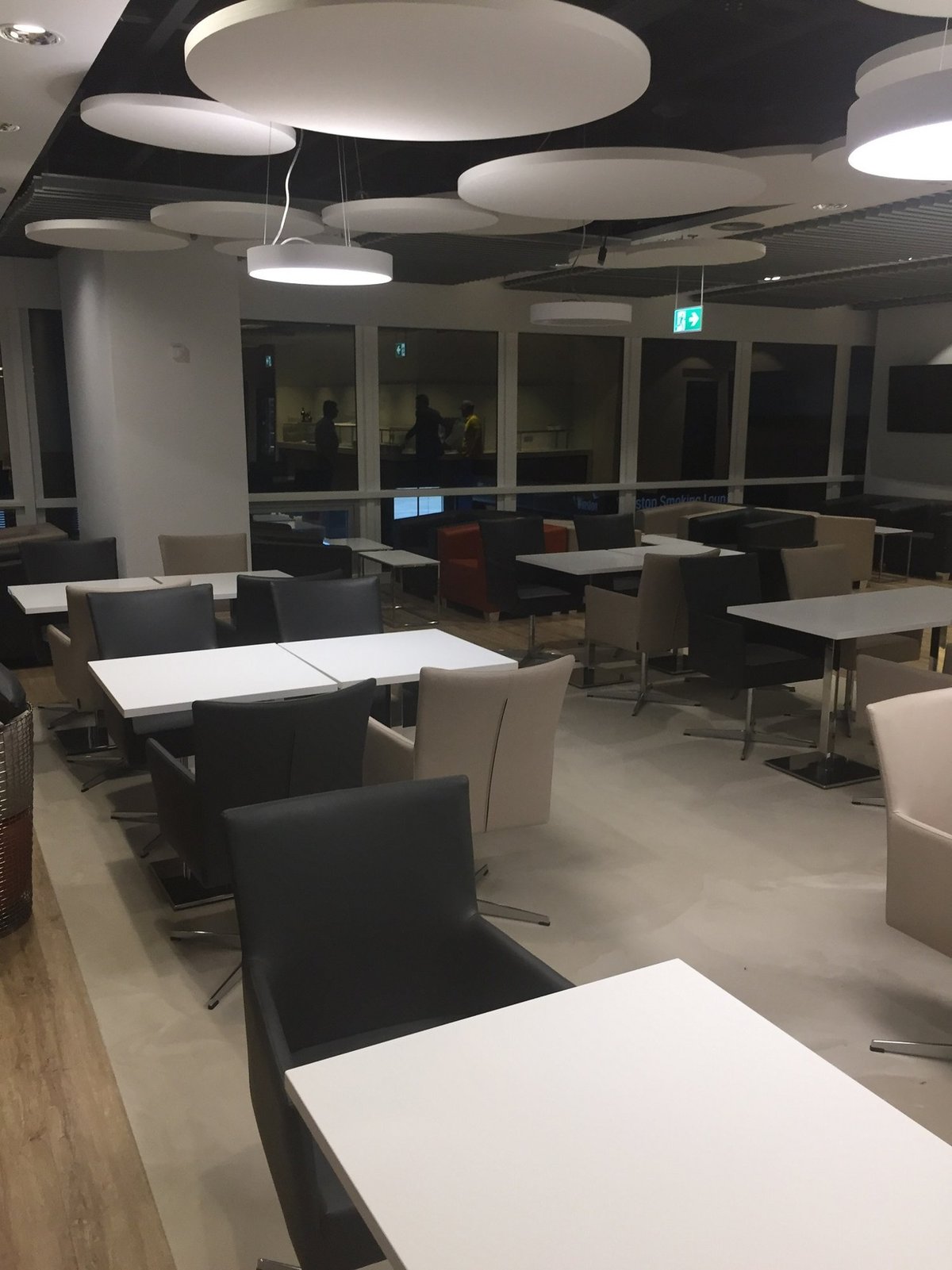 Evening view of the dining area inside Düsseldorf Airport’s Open Sky Lounge, featuring white tabletops, cushioned leather chairs in neutral tones, large modern ceiling light fixtures, and floor-to-ceiling windows overlooking the terminal.