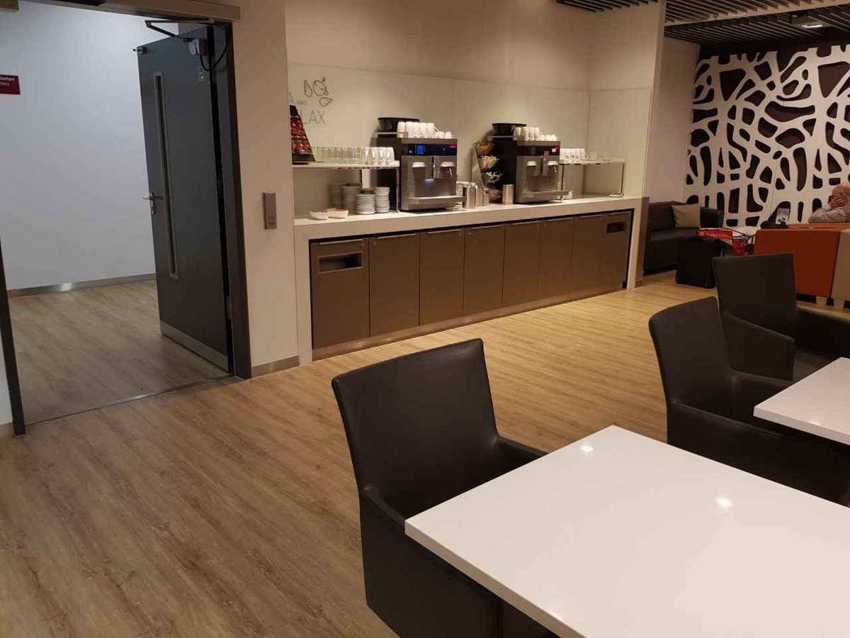 Self-service beverage station inside Düsseldorf Airport’s Open Sky Lounge, featuring sleek coffee machines, stacked cups and plates, modern brown cabinetry, and a cozy seating area with white tabletops and leather chairs on wood flooring.