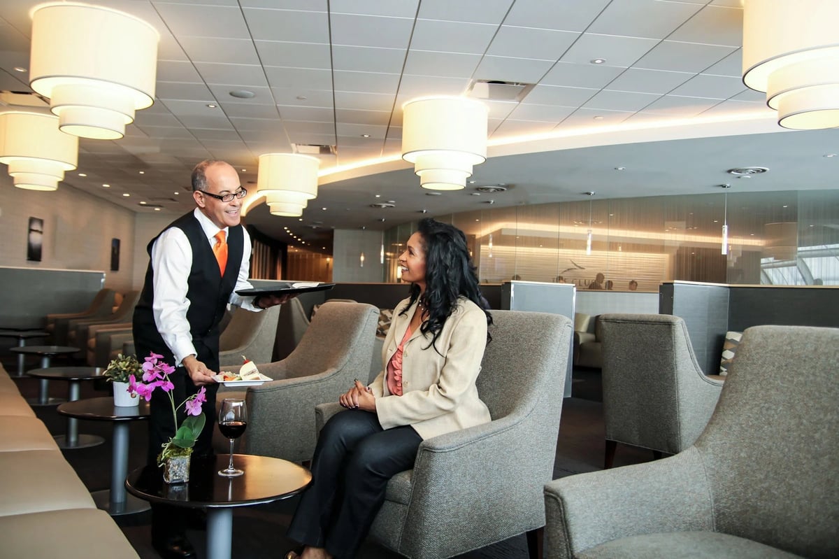 Smiling waiter serving a traveler in The Club at MCO Terminal B with stylish, comfortable seating.