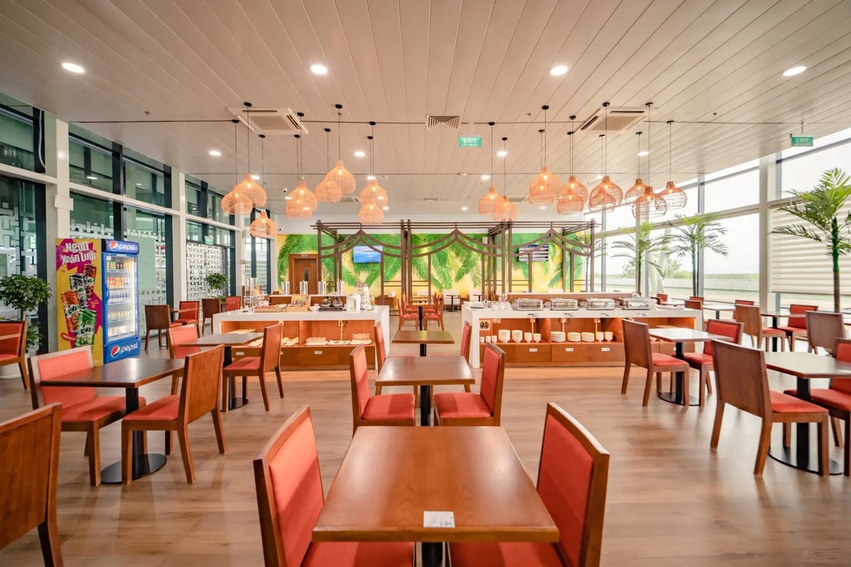 Wide view of the dining area in the SH Premium Lounge at Phu Cat Airport (UIH), showing multiple wooden tables with orange chairs, a central buffet section, tropical plant accents, and large windows.