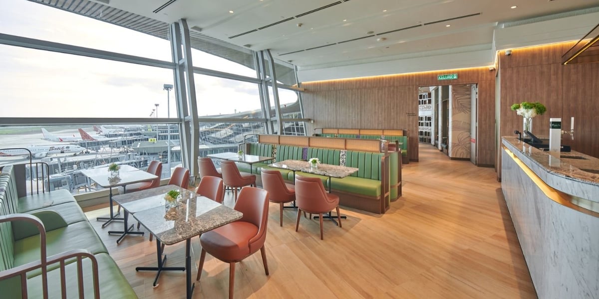 Modern dining area at the Plaza Premium First Lounge, Kuala Lumpur Airport, featuring marble tabletops, a green banquette setting, and large windows with views of aircraft at the gates.