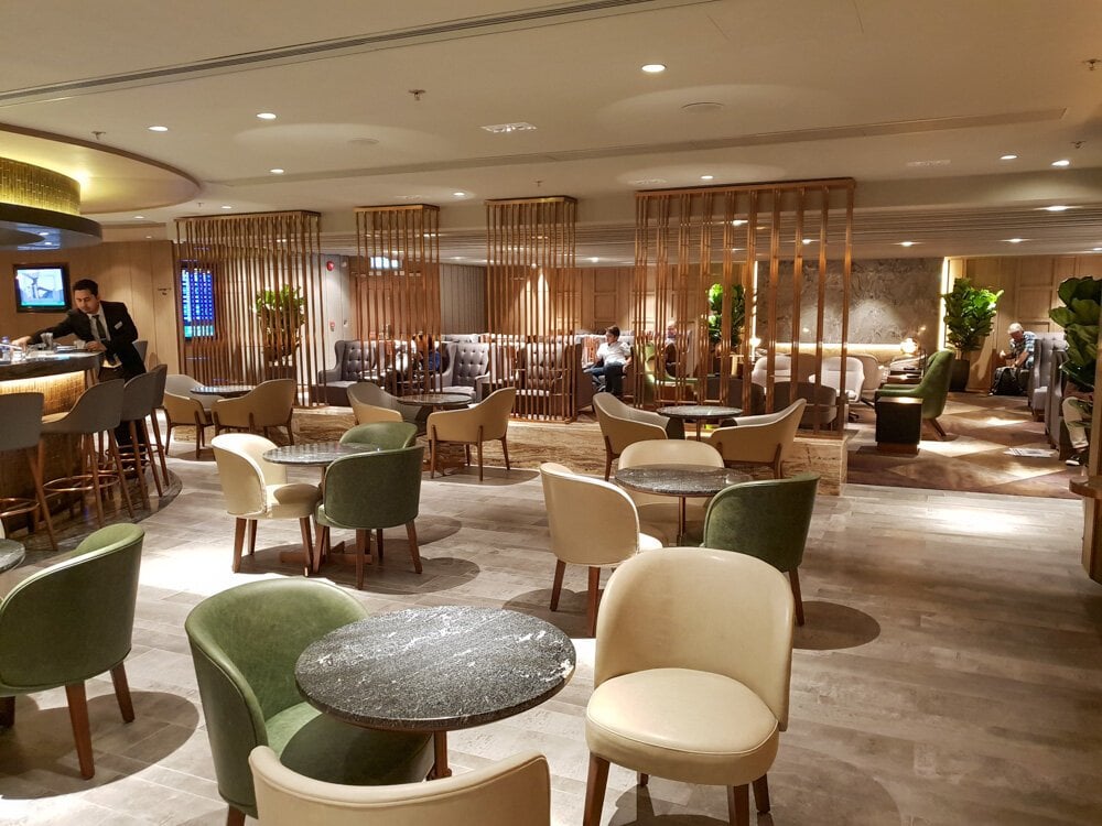 Spacious seating area at the Plaza Premium First Lounge in Hong Kong International Airport, featuring a central bar, plush armchairs in green and cream tones, and cozy private zones separated by wooden partitions.