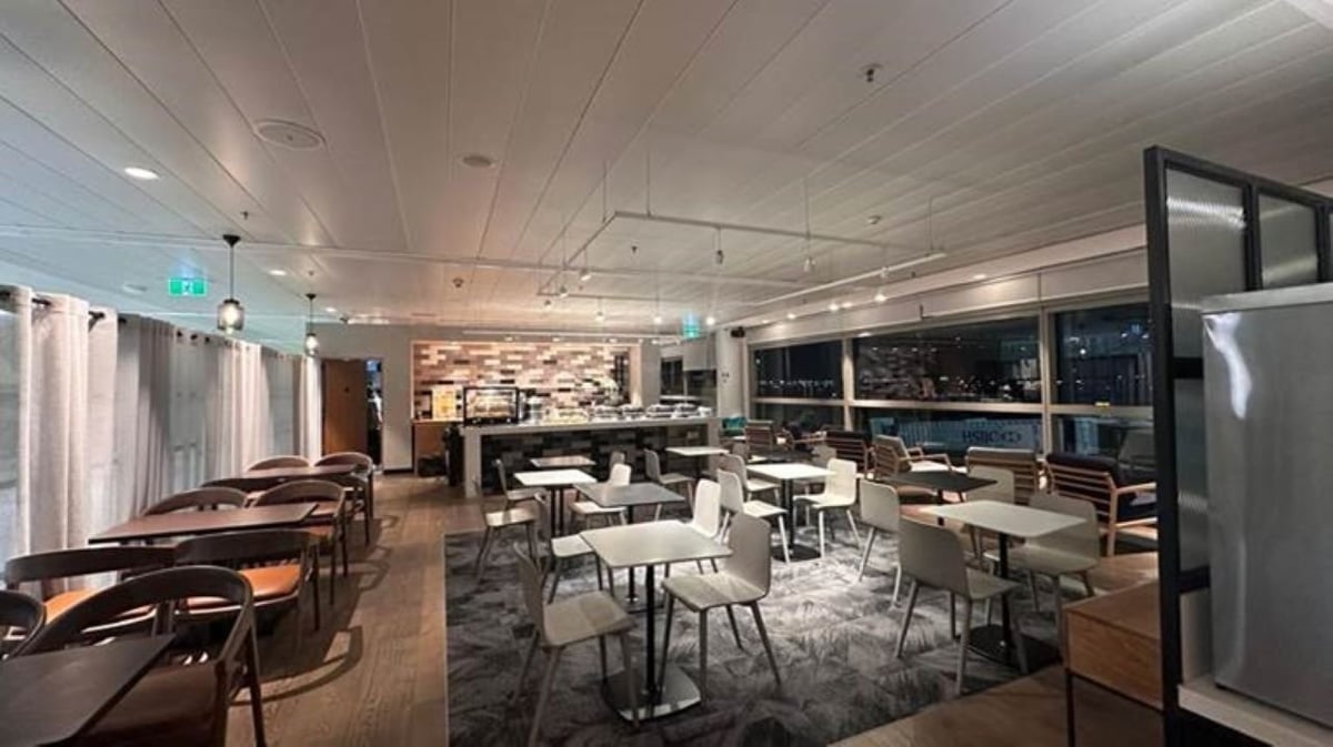 Dining area at the Plaza Premium Lounge at Brisbane Airport featuring a variety of seating options with large, floor-to-ceiling window views on either side of the room and what appears to be a buffet or bar at the back of the room.