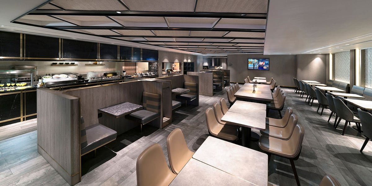 Image of the dining are of the Plaza Premium Lounge at Hong Kong Airport. Featuring a stocked buffet lining the left well and a variety of seating options including booths and dining tables. A tv is mounted on the wall at the far end of the room.