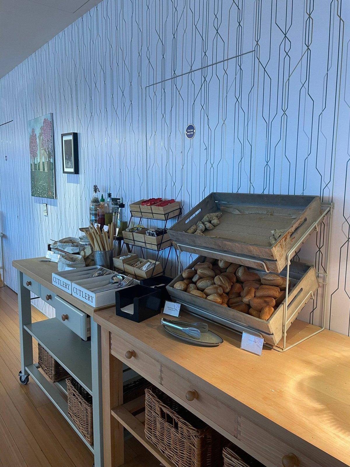 This image shows the bread station at the Plaza Premium Lounge in Gatwick Airport. The setup includes a wooden counter with baskets of freshly baked bread rolls displayed in rustic metal trays.