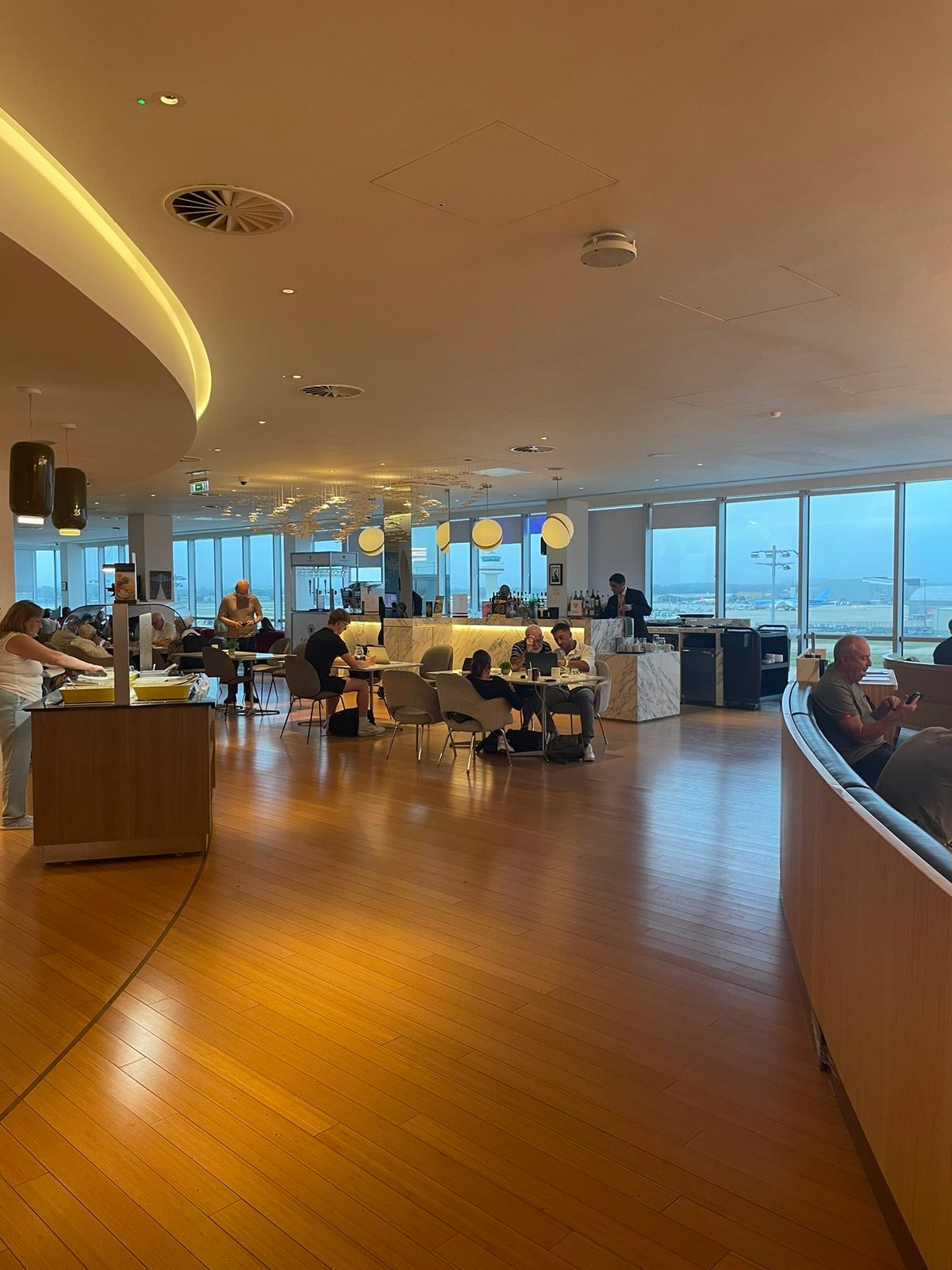 This image depicts a spacious area of the Plaza Premium Lounge at Gatwick Airport. There is sleek wooden flooring and modern decor. The lounge features a central bar area with marble accents, surrounded by tables and chairs where travellers are sat.