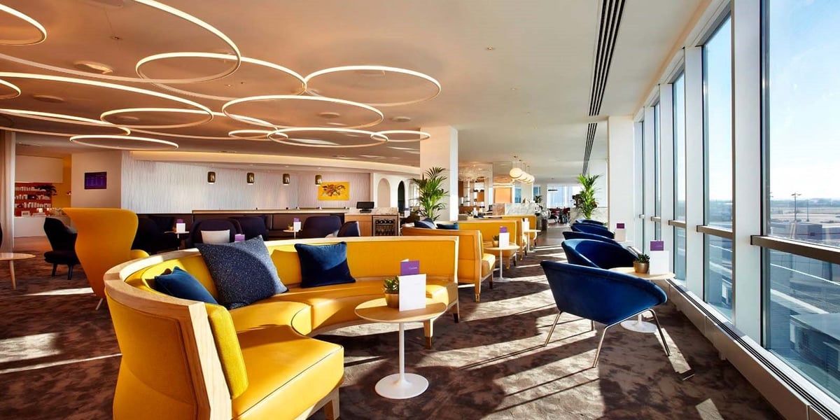 Modern airport lounge (Plaza Premium Gatwick) with circular ceiling lights, yellow sofas, blue chairs, and large windows overlooking the tarmac. Stylish bar area visible in background.