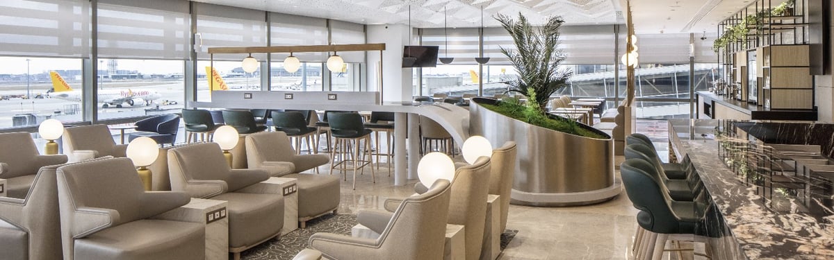 Plaza Premium Bosphorous Lounge seating area with beige armchairs with charging ports, a marbled bar and row of high stools. There is a plant centrepiece and windows overlooking the runway to the left.