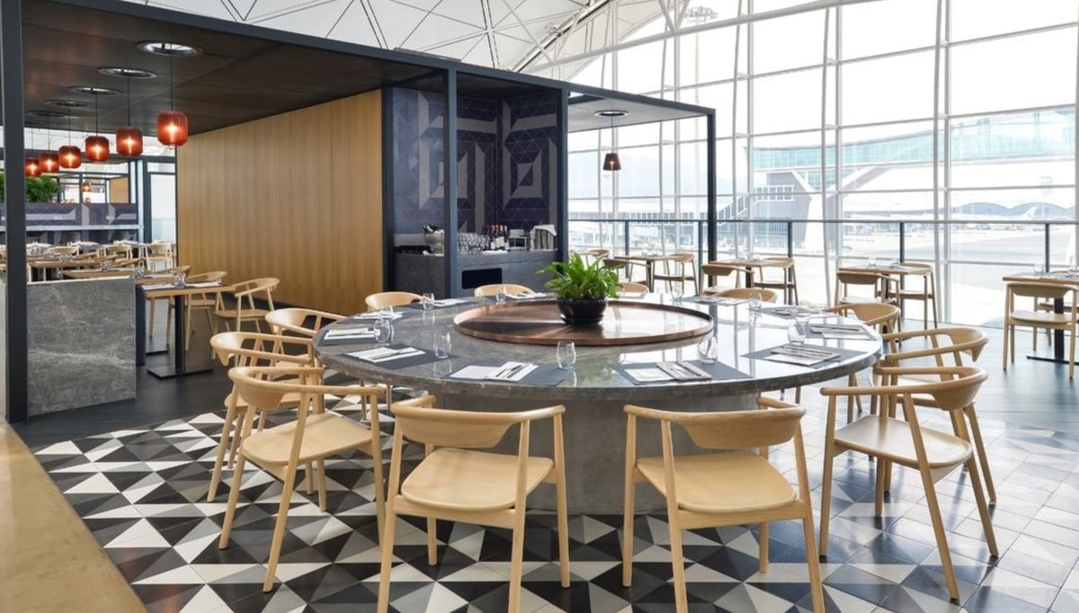 Spacious dining area at the Qantas Hong Kong Lounge, featuring a large communal stone table surrounded by light wooden chairs, geometric tiled flooring, and floor-to-ceiling windows with panoramic airport runway views.