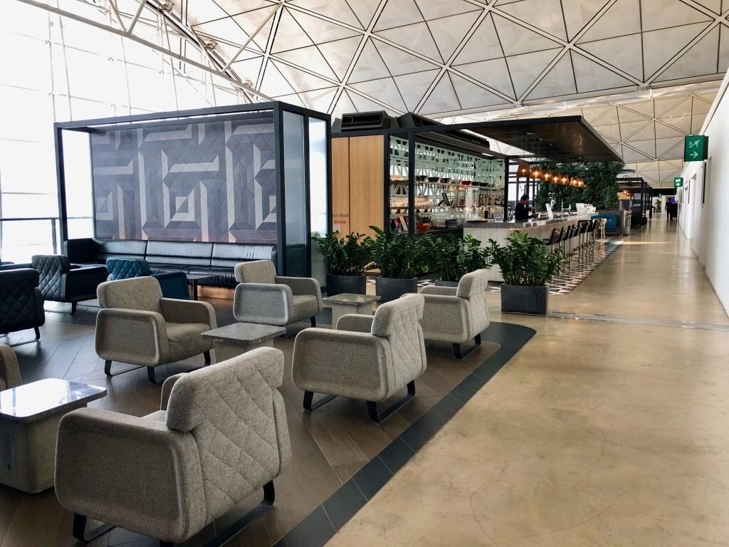Qantas International lounge at Hong Kong Airport featuring partitioned zones at the lounge, one seating area with plush grey armchairs, and another zone being the bar which is partitioned by greenery in pots.