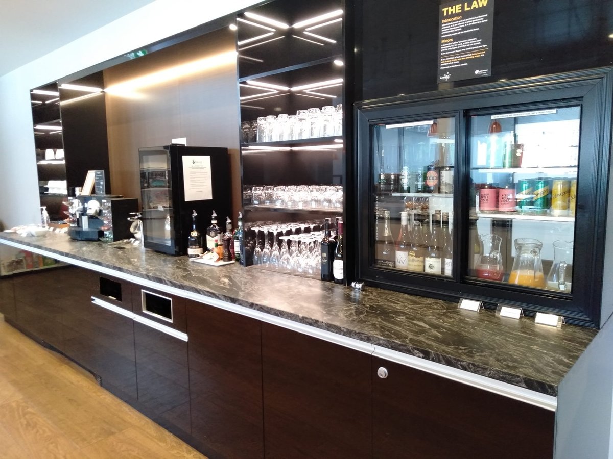 Beverage section of the Manaia Lounge at Queenstown Airport. There is a mini fridge to the right on marbled countertops displaying alcoholic drinks, juices and soft drinks. Beverage glasses are in the middle, and a coffee machine is to the far left.