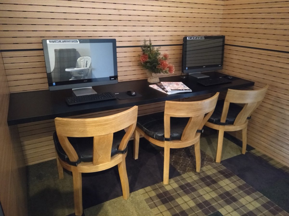 Workstation at the Manaia Lounge at Queenstown airport. The station features wooden chairs with leather seating and two computers. There is a selection of magazines between the computers and decorative greenery.
