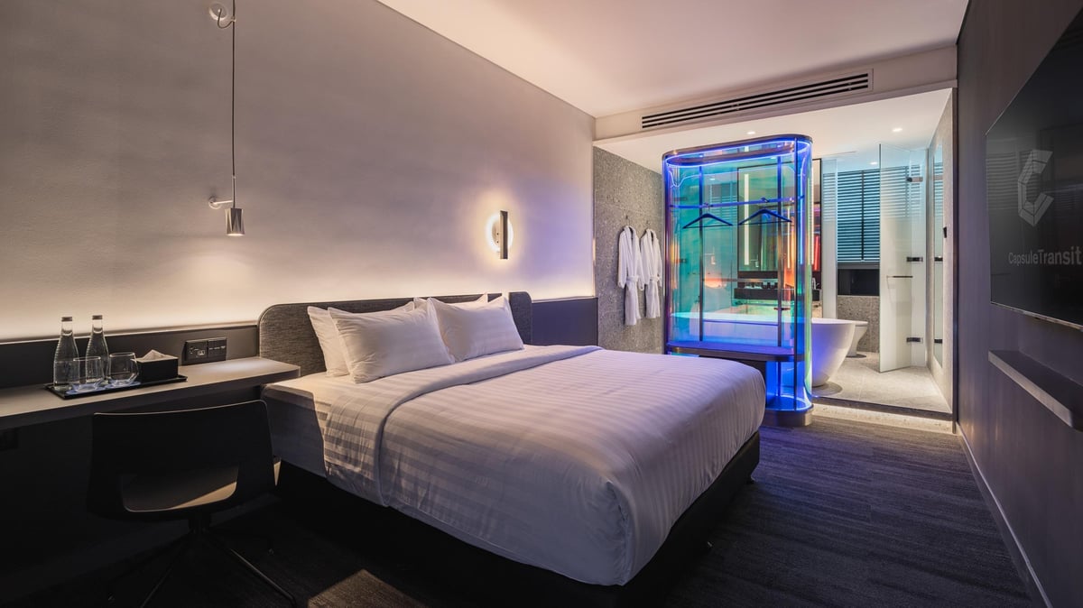 Sleek and modern guest room at Capsule Transit MAX, KLIA Terminal 2, featuring a large bed with crisp white linens, a compact work desk, ambient lighting, and an ensuite bathroom with glass walls, a freestanding tub, and a blue-lit wardrobe.