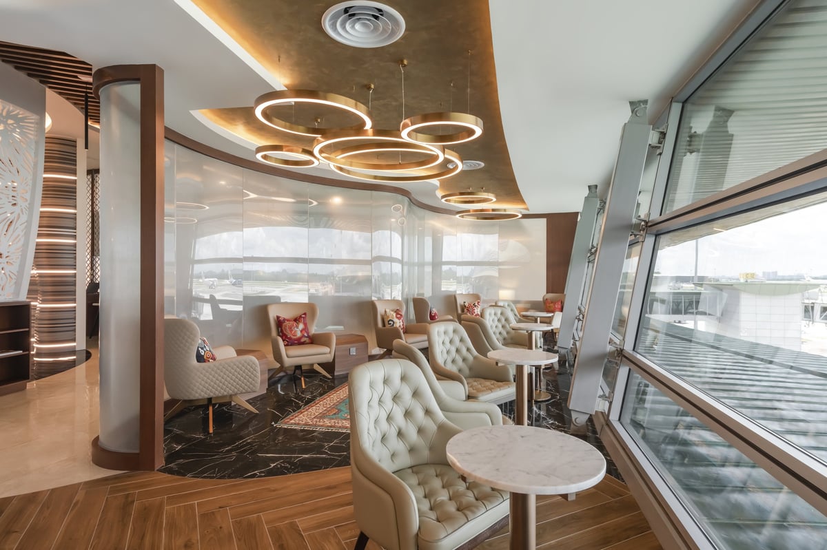 Relaxing tarmac-view seating area at the Travel Club Lounge in KLIA’s Satellite Terminal, featuring tufted leather chairs, small marble tables, large slanted windows, and sculptural ring lighting overhead.
