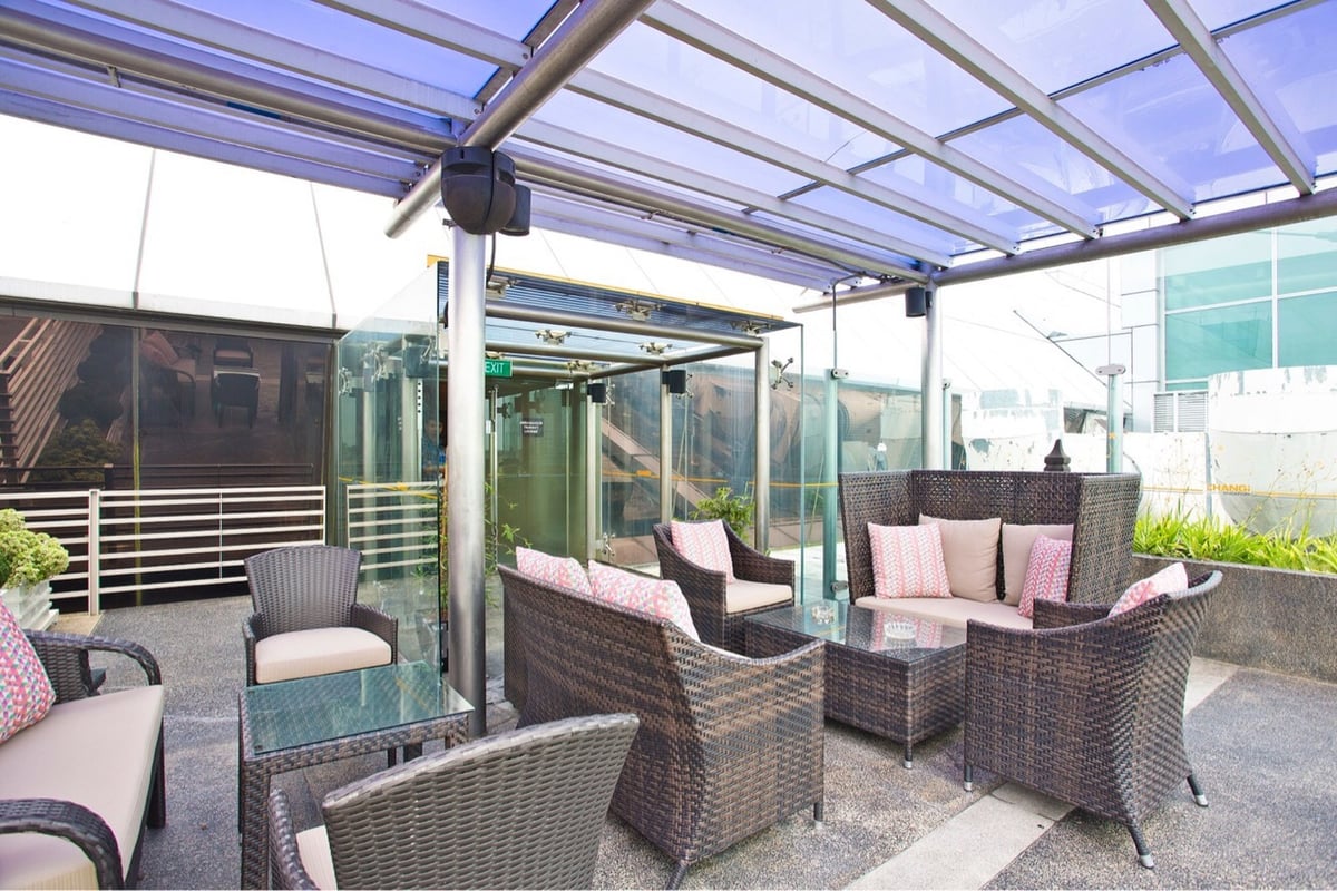 Outdoor seating area at Ambassador Transit Lounge, Changi Airport Terminal 2, featuring wicker armchairs and sofas with pastel cushions under a glass canopy, creating a relaxed, airy space for travelers.
