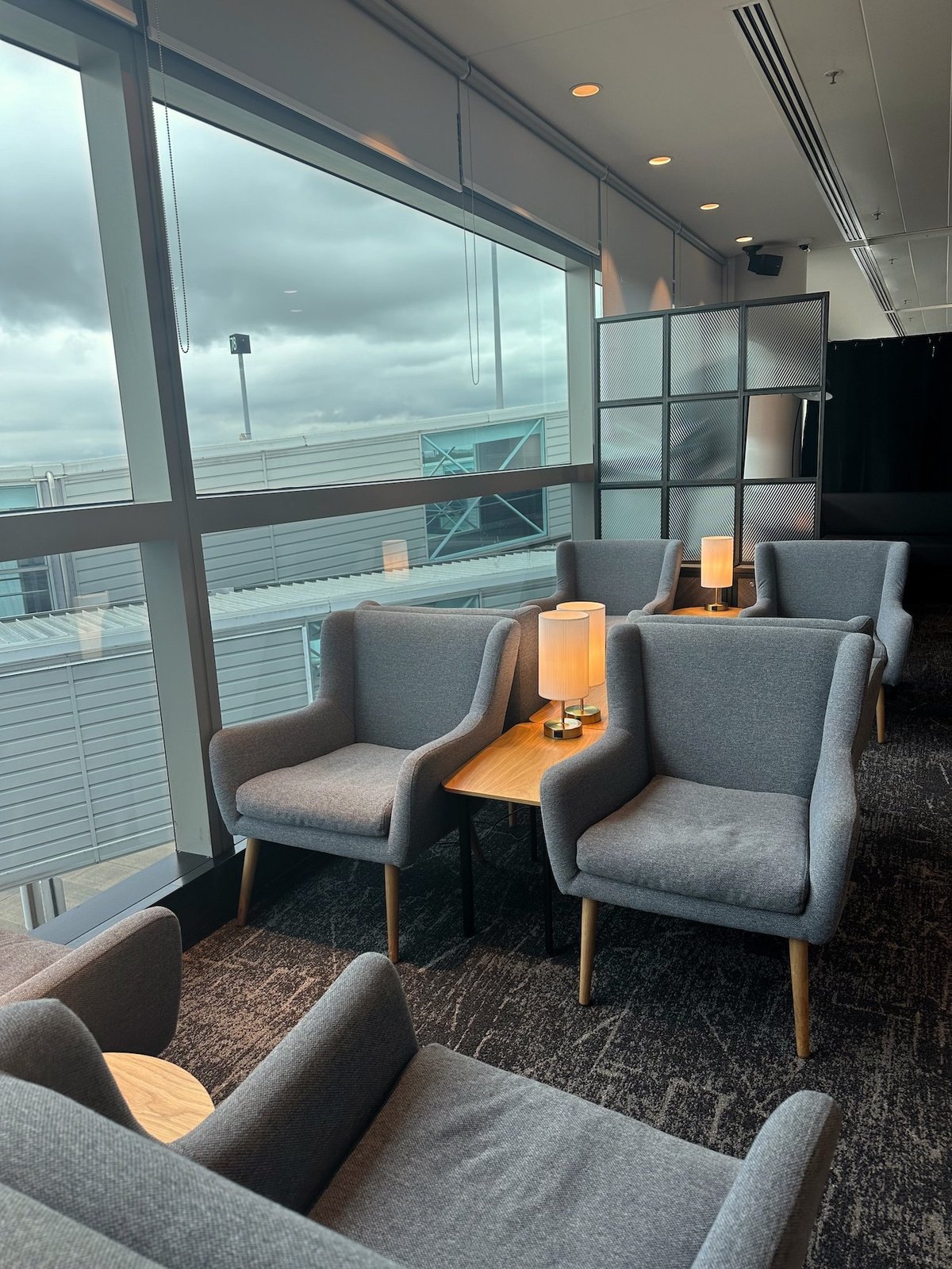 Grey plush seats next to floor to ceiling windows at the Plaza Premium Lounge at Brisbane Airport. Between the seats are low wooden tabletops with contemporary lamps illuminating the space.