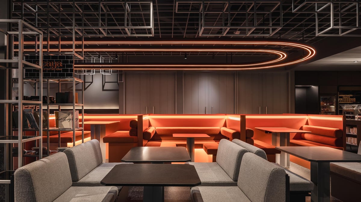 Capsule Transit MAX lounge seating area at KLIA Terminal 2, featuring a mix of cozy orange booth seating and sleek grey upholstered chairs, illuminated by soft linear ceiling lights and modern geometric design elements.