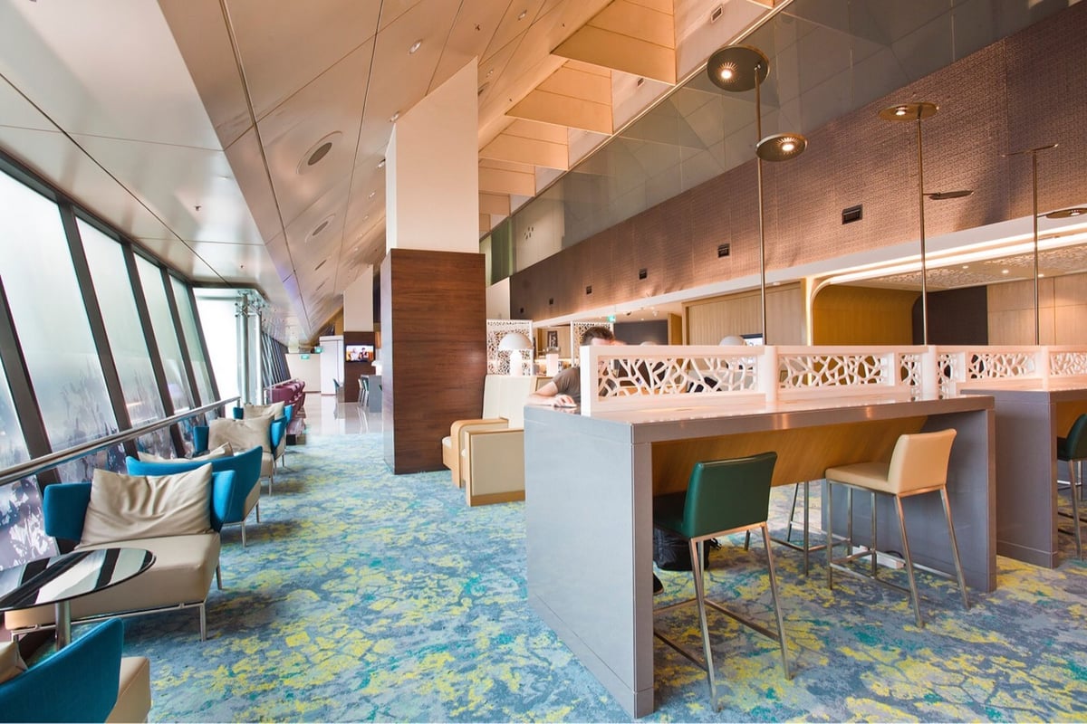 Stylish seating area in Ambassador Transit Lounge at Changi Airport Terminal 2, featuring modern armchairs, high work tables, colorful patterned carpet, and large slanted windows with natural light.