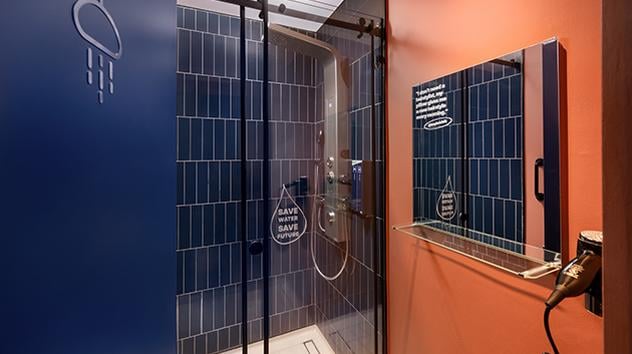 Shower at the Kepler Club at the Main Terminal Building, Kuala Lumpur Airport. There is a shower in the image, a mirror and hairdryer attached to the wall, the wall has a blue and rust colour scheme.