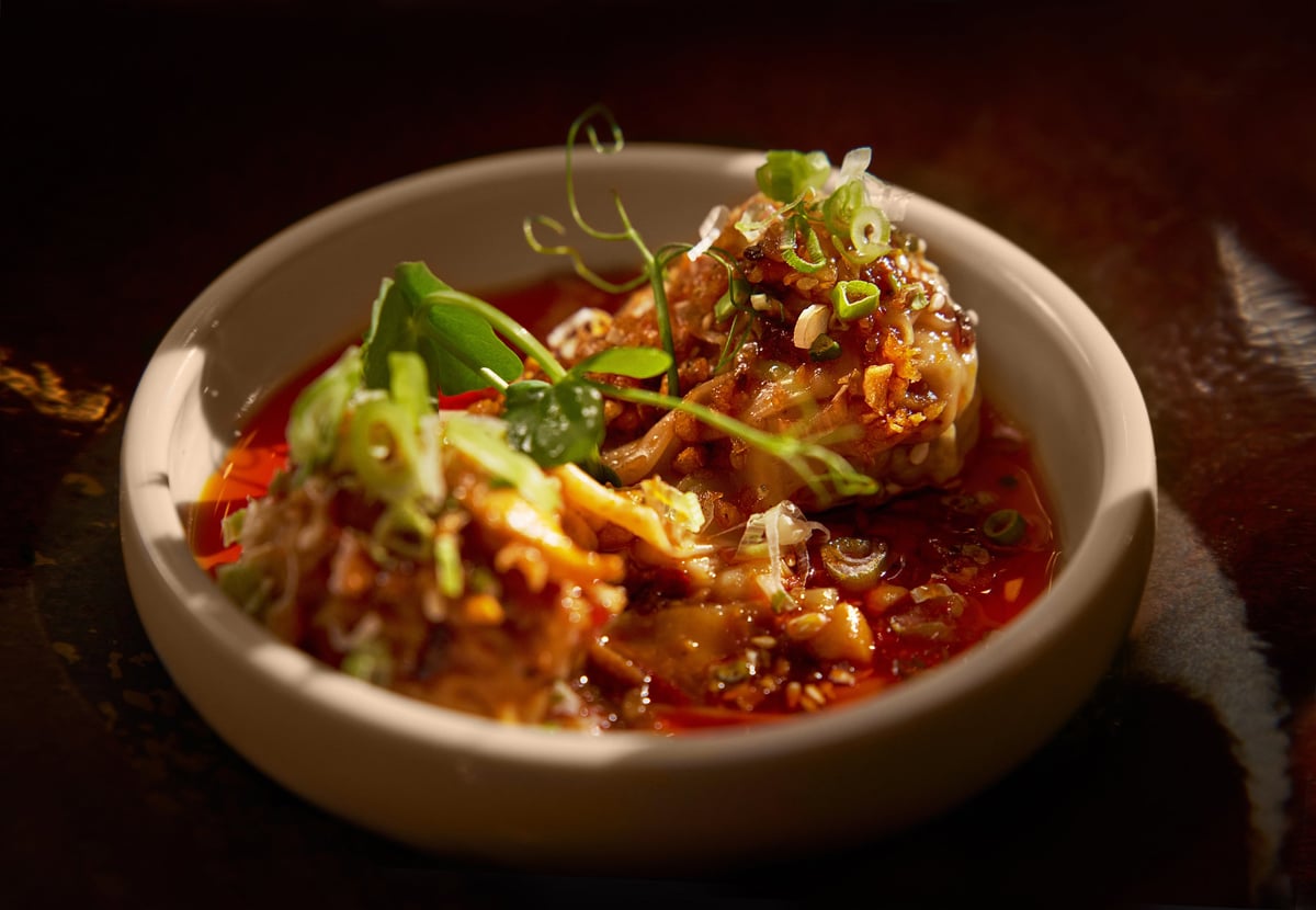 A close-up of hand-wrapped Sichuan pork dumplings served at The Pier First Class Lounge in Hong Kong International Airport, presented in a shallow bowl with spicy chili oil, garnished with chopped scallions, crispy garlic, and microgreens.