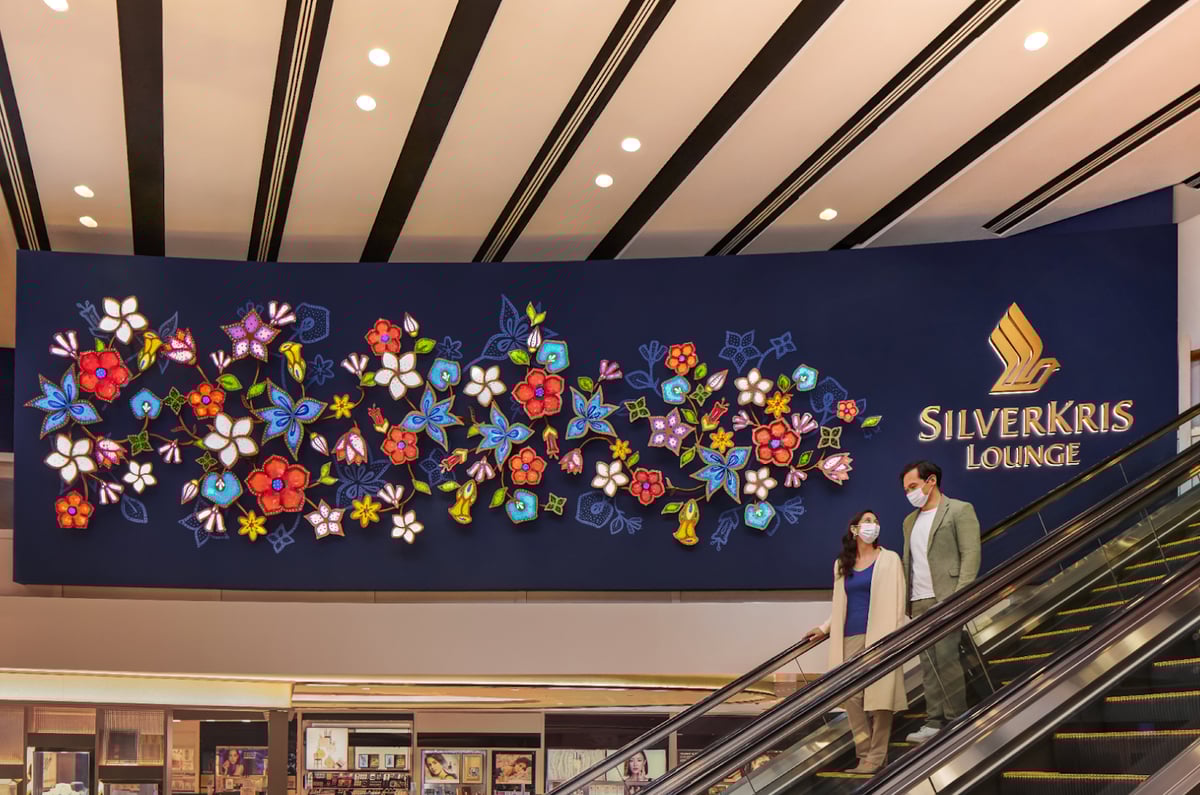 Colorful floral wall art installation and SilverKris Lounge signage at Changi Airport, featuring two travelers on an escalator wearing face masks.