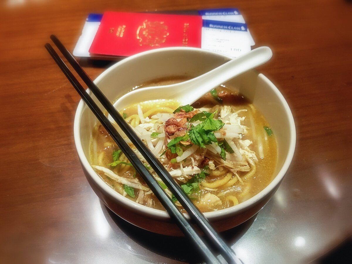 Traditional bowl of Singapore Laksa noodles served at the SilverKris Lounge in Singapore Changi Airport, photographed with chopsticks, a spoon, and a passport in the background.