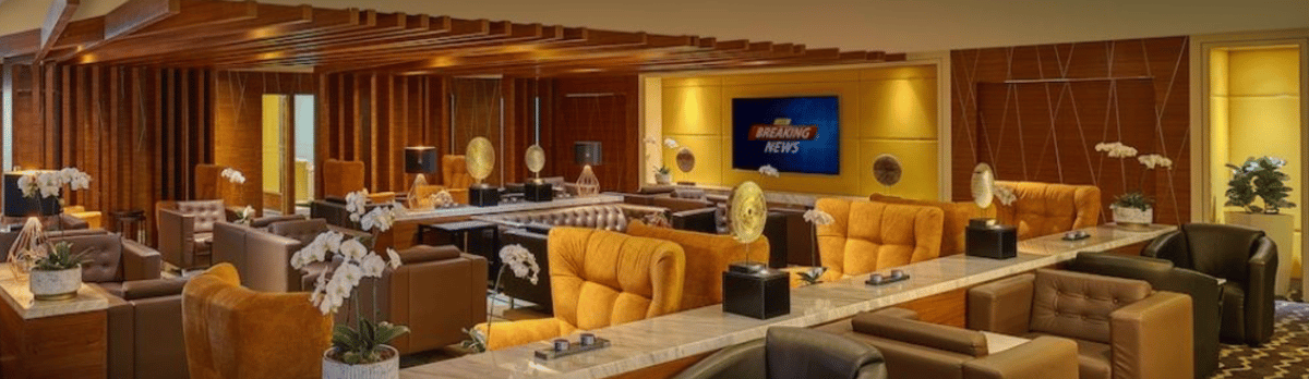 Warm and modern seating area inside the Sky Suite Airport Lounge at KLIA Terminal 2, featuring plush orange and brown armchairs, marble-topped tables, soft lighting, decorative art pieces, and a flat-screen TV on a golden accent wall.