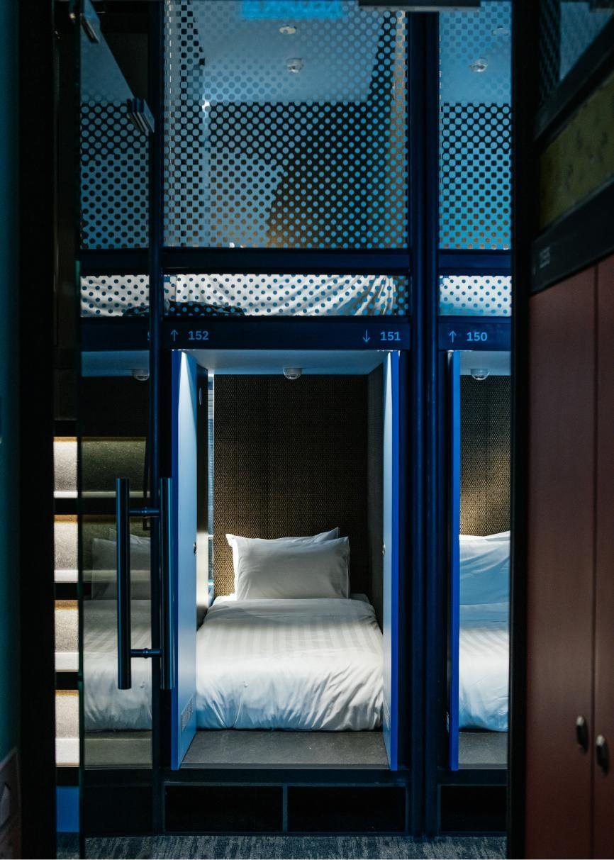 Sleek single sleeping pod at CapsuleTransit KLIA Terminal 2 (Airside), featuring a neatly made bed with white linens enclosed in a futuristic blue and black cubicle-style design, with numbered labels and a patterned privacy screen above.