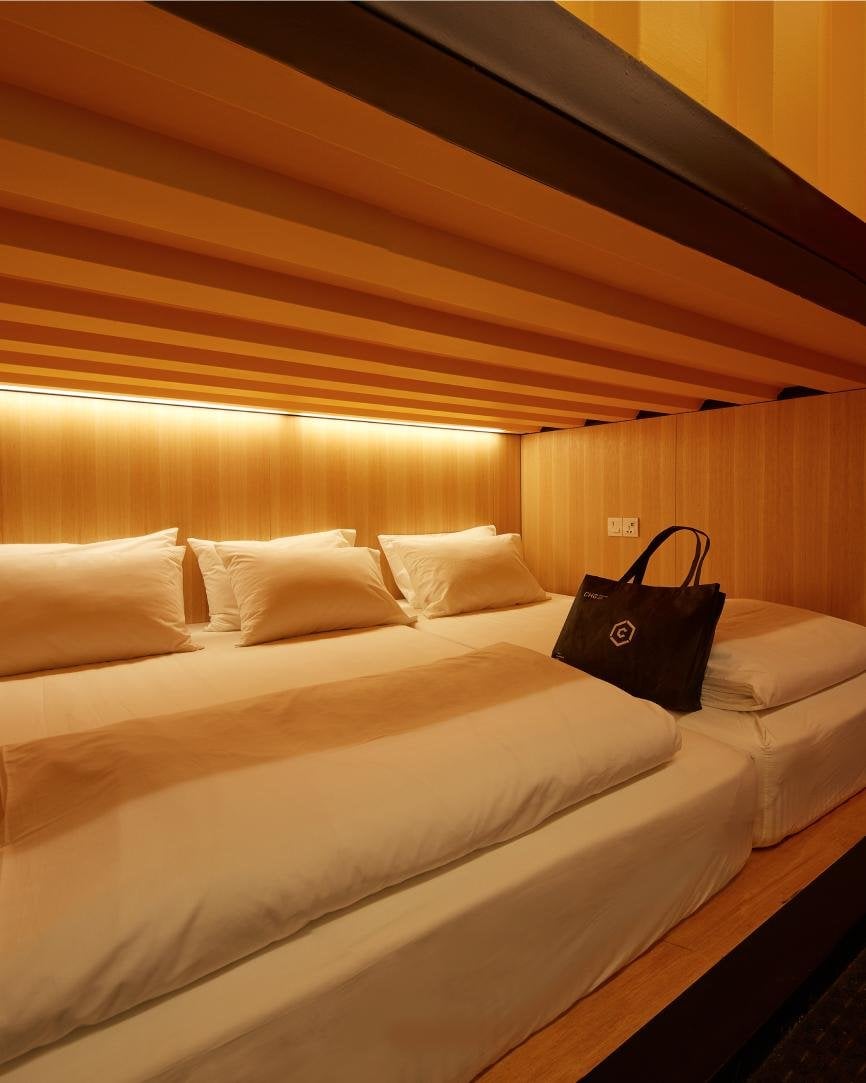 Warmly lit suite at CapsuleTransit KLIA Terminal 2 featuring a spacious double bed with crisp white linens and pillows, set against smooth wood-paneled walls with a cozy, minimalist aesthetic. A branded black tote bag rests on the bed.