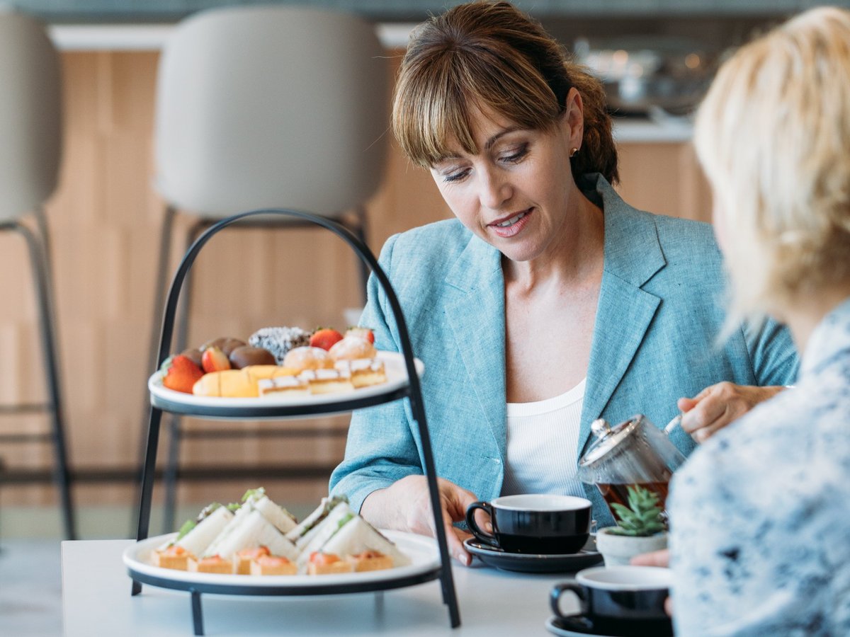 Plaza Premium Lounge at Sydney Airport image showcasing guests pouring coffee and a selection of food and snacks are positioned on the table. Food includes sandwiches, lamingtons, strawberries and various sweets.