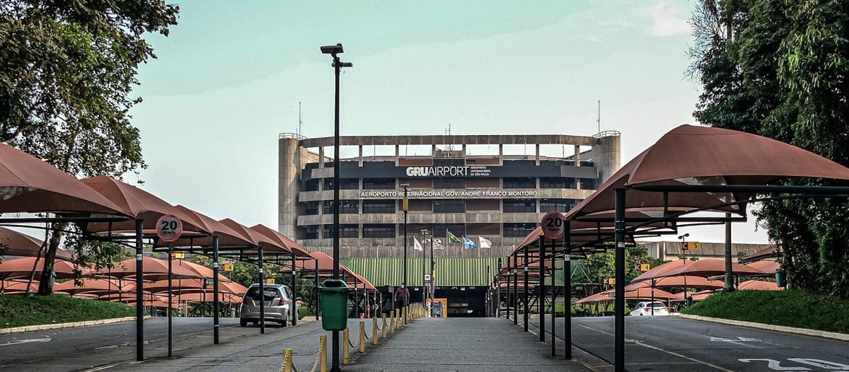Exterior of GRU Airport Terminal 2 in São Paulo, Brazil, with covered walkways, parked cars, and flags near the entrance