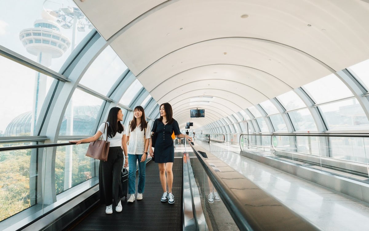 Travellers walking on travellator inside glass walkway at Changi Airport with control tower view in background.