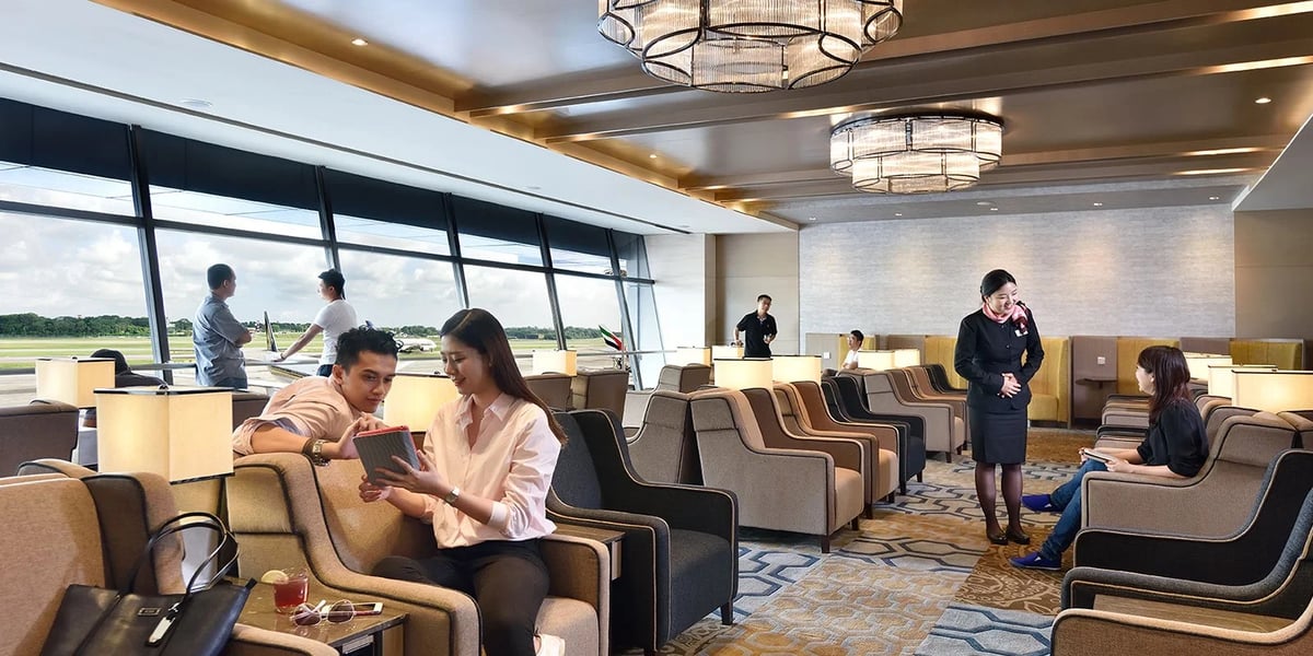 Seating area of the Plaza Premium Lounge at Singapore Changi Airport. Colour palette is neutrals with bright ceiling light fixtures. People are sitting and standing in various locations around the lounge.