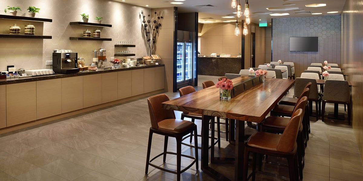 Dining and buffet area in the Plaza Premium Lounge at Changi Airport. A elongated wooden dining table rests in the foreground, while to the left is the buffet area with decorative potted plants, a coffee machine, utensils, glasses and a fridge.