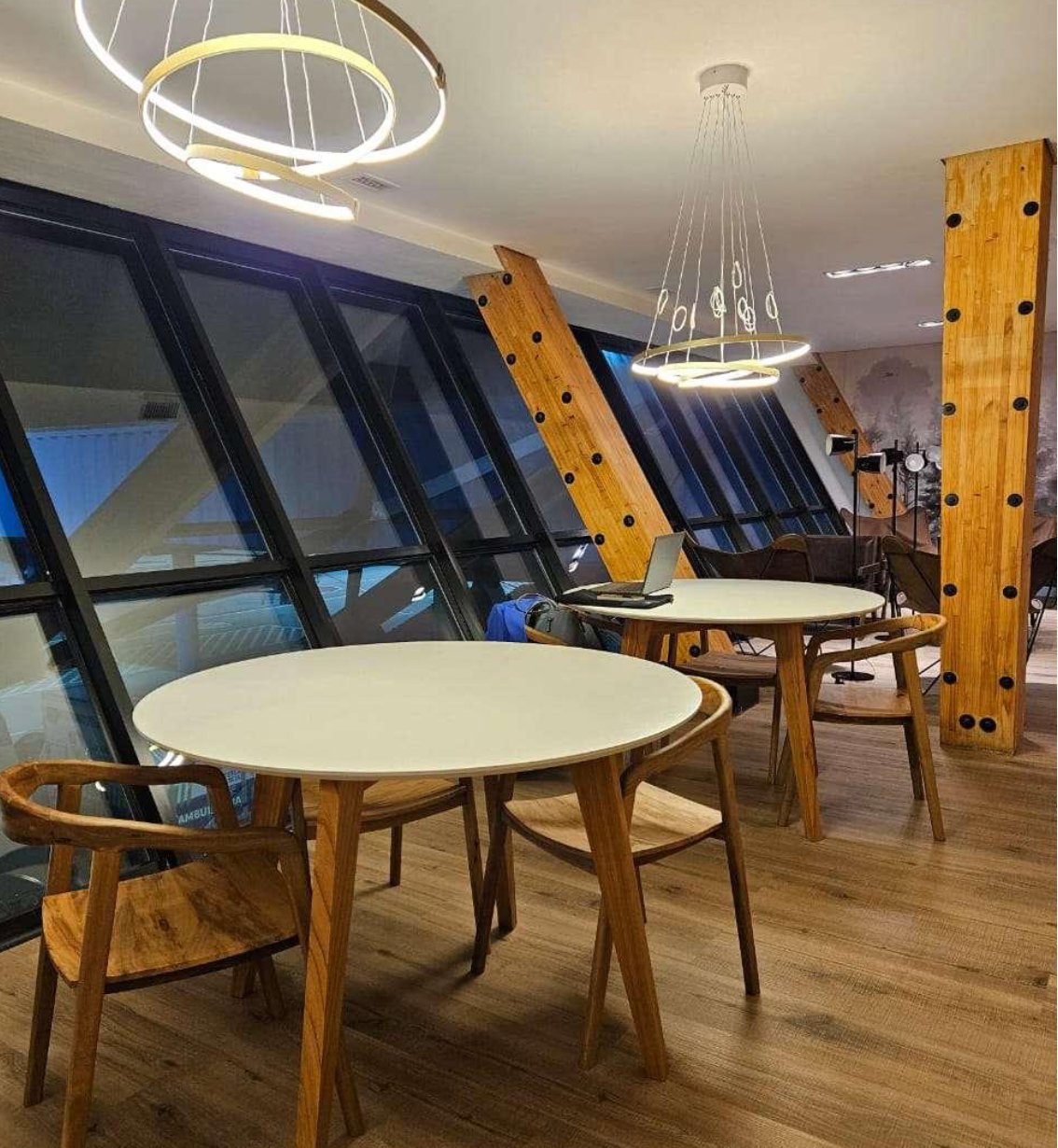 Work and dining area in the W Lounge at Ushuaia International Airport, featuring round white tables, wooden chairs, angled floor-to-ceiling windows, and modern ring pendant lighting with exposed wooden beams.