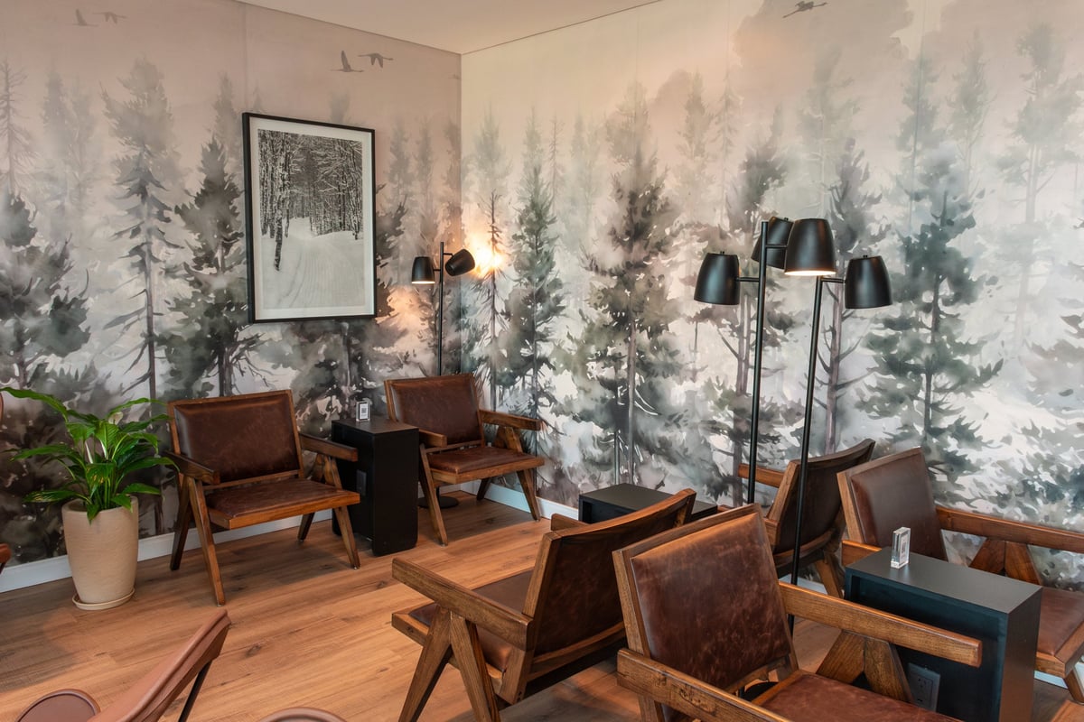 Cozy seating corner in the W Lounge at Ushuaia Airport, featuring rustic wood and leather armchairs, matte black floor lamps, and a serene forest-themed mural with a snowy path photograph on the wall.