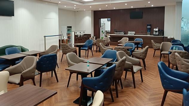 The Lounge at Ahmedabad Airport (AMD) Terminal 2 featuring modern seating with blue and beige chairs, wooden tables, and a staffed food and beverage counter in the background.