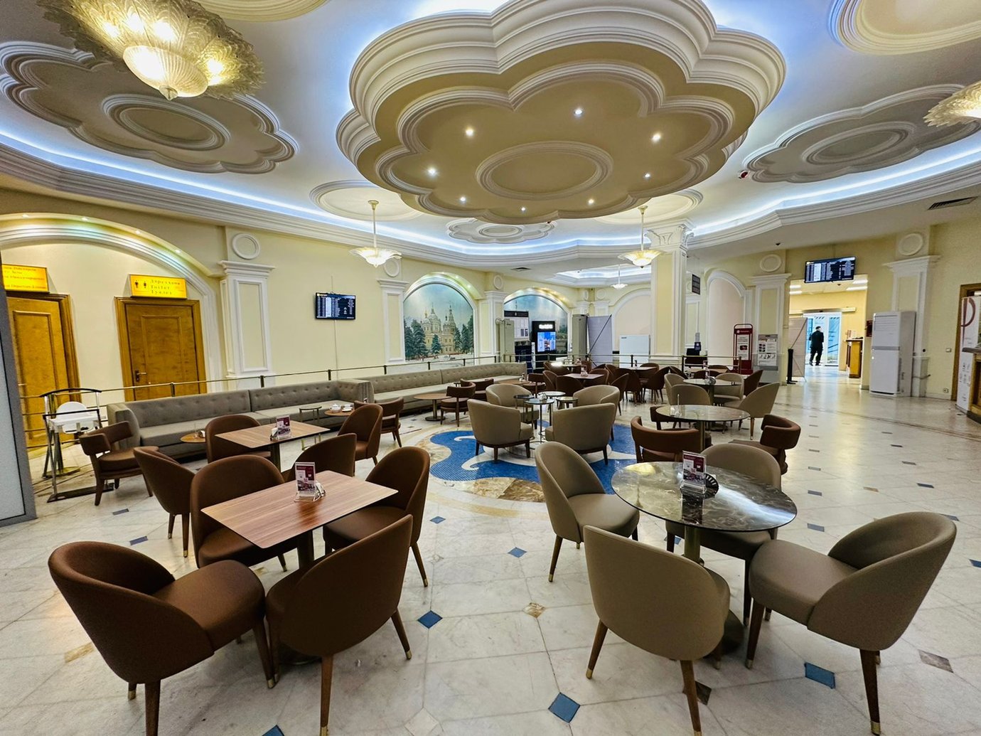 seating area inside the Primeclass Lounge at Almaty International Airport, featuring tan and brown chairs, marble flooring, ornate white ceilings with recessed lighting, and classic murals on the walls.
