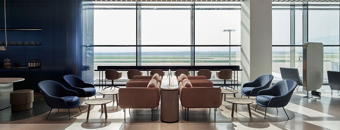 Elegant seating area in the Aegean Lounge at Athens Airport, showcasing a stylish blend of modern and classic design elements. There are armchairs in deep blue and rose colours, complemented by small marble roundtables. A panoramic window at the back Elegant seating area in the Aegean Lounge at Athens Airport, showcasing a stylish blend of modern and classic design elements. There are armchairs in deep blue and rose colours, complemented by small marble roundtables. A panoramic window at the back