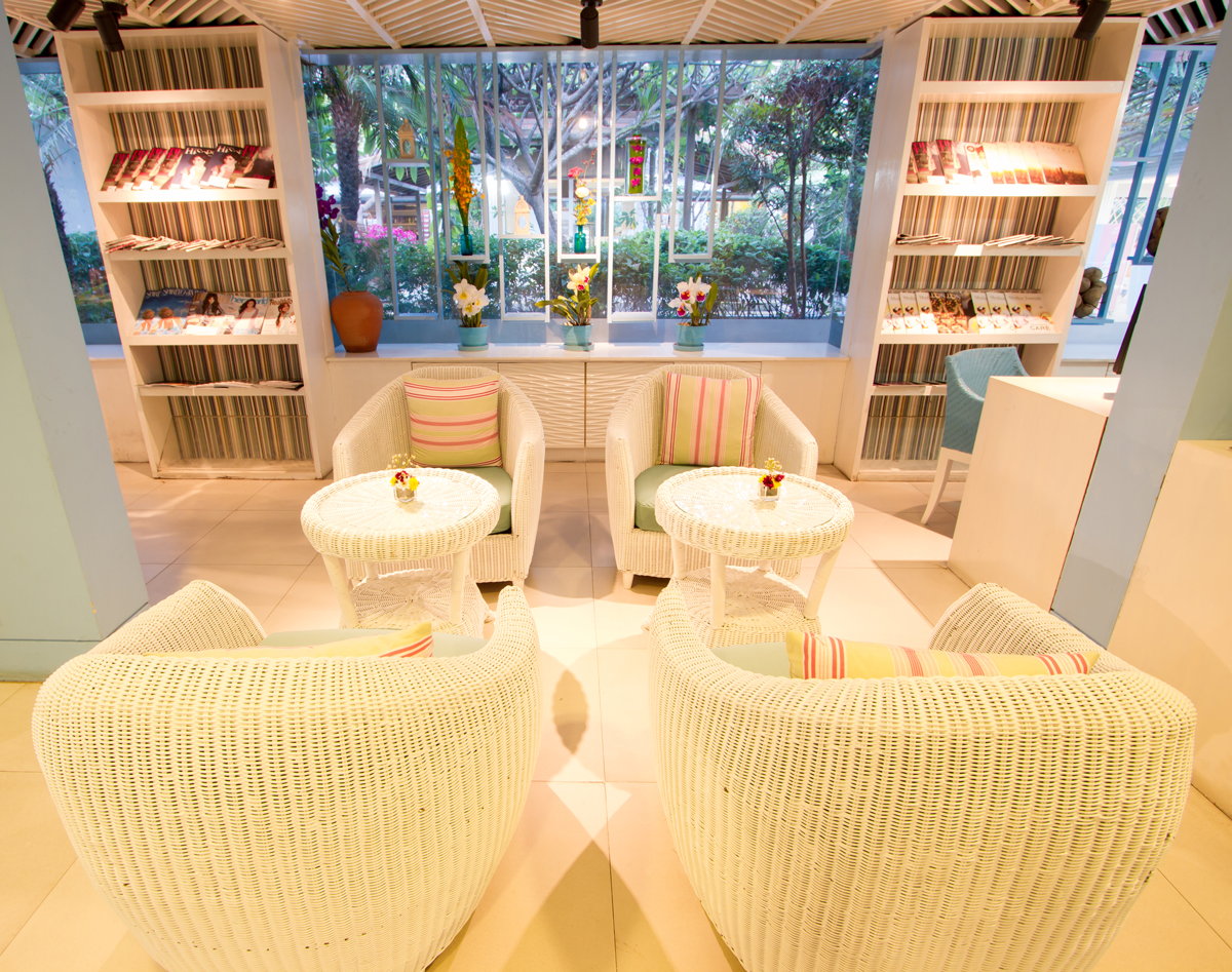 Bright lounge seating area with white wicker chairs, small round tables, magazine shelves, and a window overlooking a lush garden.