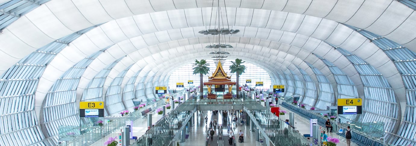 Interior view of Suvarnabhumi Airport in Bangkok featuring the distinctive arched glass ceiling, Thai-style pavilion, palm trees, and passengers walking through the bright terminal. Interior view of Suvarnabhumi Airport in Bangkok featuring the distinctive arched glass ceiling, Thai-style pavilion, palm trees, and passengers walking through the bright terminal.
