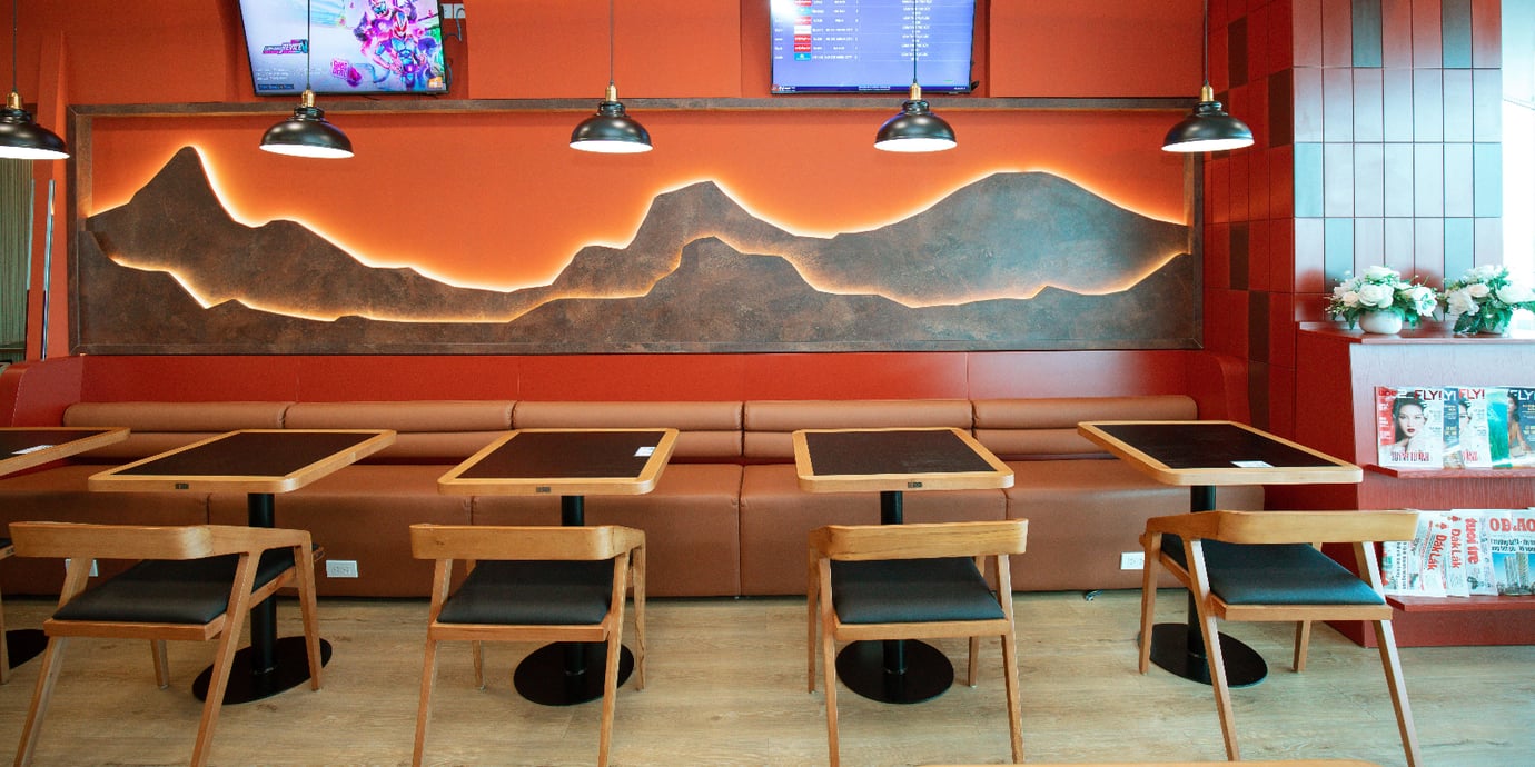 SH Premium Lounge Buon Ma Thuot Airport (BMV) with a cozy dining-style seating area, featuring booth benches, wooden tables and chairs, warm orange lighting, and modern décor.