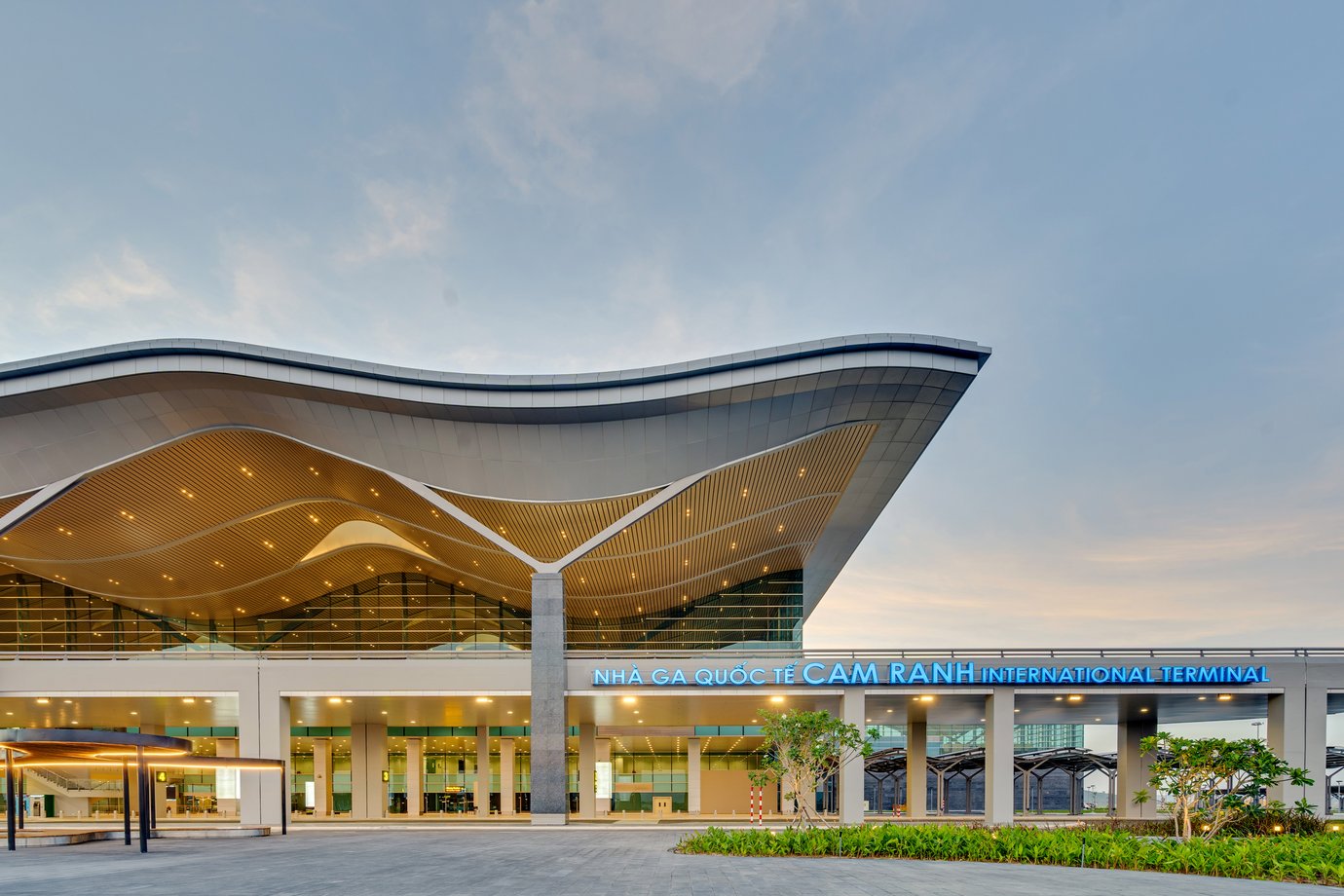 Exterior view of Cam Ranh International Airport terminal in Vietnam with modern wave-shaped roof design at sunset.