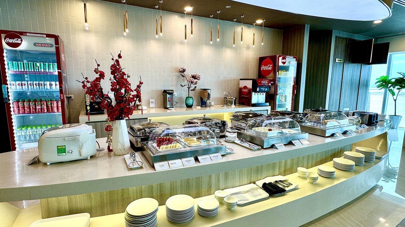 Buffet counter at Sun Coast Business Lounge in Cam Ranh International Airport Terminal 2, featuring hot dishes, fresh fruit, desserts, beverages, and Coca-Cola drink station.