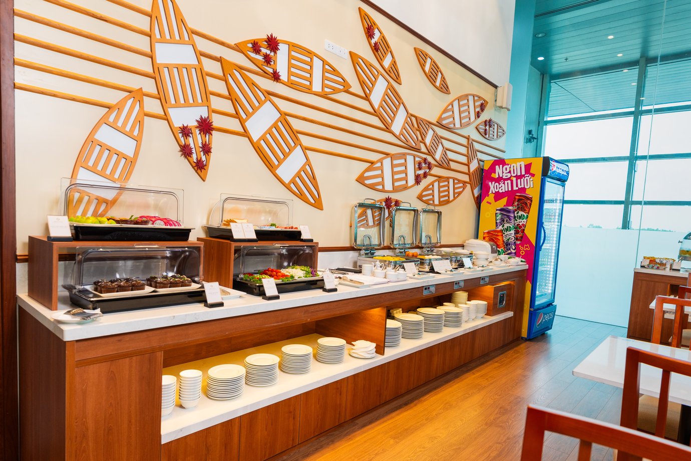 SH Premium Lounge buffet at Can Tho International Airport (VCA), featuring a bright self-serve spread of snacks, desserts, and beverages, with warm wood finishes, decorative wall art, and a relaxed dining area.
