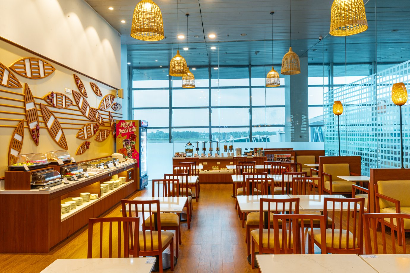 SH Premium Lounge dining area at Can Tho International Airport (VCA), showcasing a spacious, light-filled seating layout with large runway-facing windows, warm wicker pendant lighting, wood-and-upholstered tables, and an easy-access buffet counter