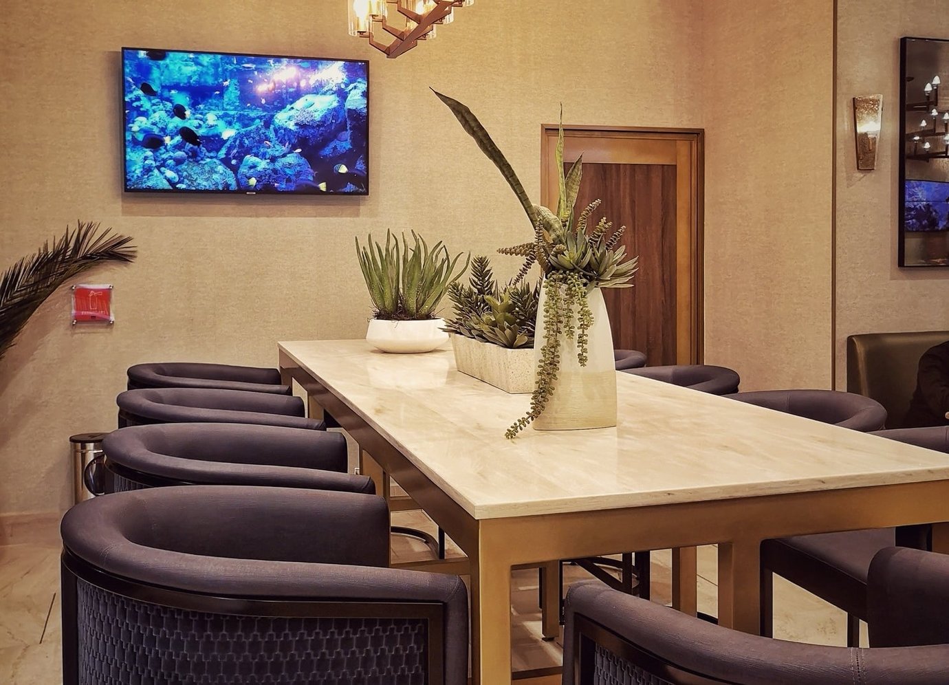 A communal high table in the Mera Business Lounge Cancun surrounded by dark upholstered chairs, with potted plants arranged along the tabletop. A communal high table in the Mera Business Lounge Cancun surrounded by dark upholstered chairs, with potted plants arranged along the tabletop.